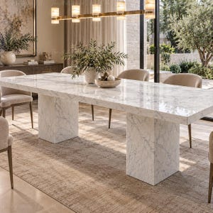 May include: A rectangular dining table featuring a polished white marble top with grey veining and two matching thick, block-style marble pedestal bases. The table is surrounded by several upholstered chairs with light beige fabric seats.