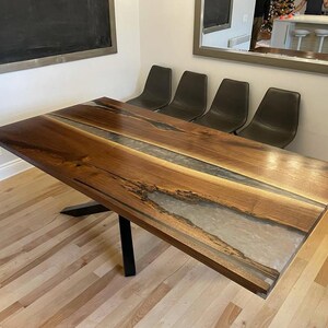 May include: A rectangular wooden dining table with a dark walnut finish and a clear epoxy river down the center. The table has a black metal base and is surrounded by four black chairs.