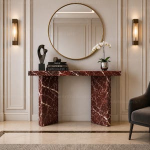 May include: A rectangular console table made of deep red marble with white veining, featuring two thick slab legs. On top sits a stack of three books titled Vogue and Kate Moss, a dark abstract sculpture, and a white orchid in a silver pot.
