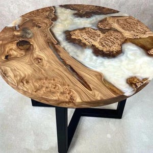 May include: Circular wooden table with a distinctive design. The tabletop showcases a light brown wood grain with a white and brown resin river. The table has a black metal base. The table is approximately 91 cm in diameter.