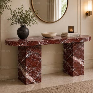 Può includere: Un tavolo console in marmo rosso intenso con un piano ovale liscio e due gambe a blocco rettangolari. La superficie presenta venature bianche prominenti su tutta la pietra. Il tavolo è posizionato contro una parete, sostenendo un vaso scuro e una ciotola.