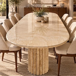 May include: An oval dining table with a polished beige travertine stone top and a fluted cylindrical pedestal base. The table is surrounded by eight upholstered chairs with dark wooden legs, featuring a smooth, light-coloured fabric finish.