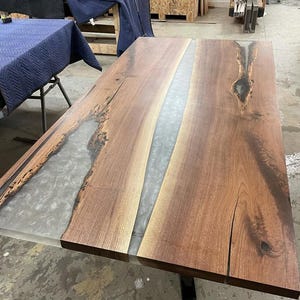 May include: A rectangular wooden table with a river of gray epoxy resin running down the center. The wood has a rich brown color with natural grain patterns and knots. The table is a modern design.