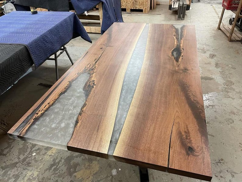 May include: A rectangular wooden table with a river of gray epoxy resin running down the center. The wood has a rich brown color with natural grain patterns and knots. The table is a modern design.