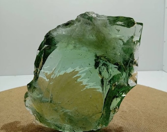 May include: A large, translucent green glass rock with a rough, irregular surface. The rock is sitting on a brown, textured surface.