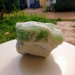 Puede incluir: Un gran cristal natural de forma irregular, verde y blanco. El cristal tiene una superficie rugosa y texturizada y está sentado sobre una superficie blanca.