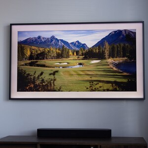 Pode incluir: Fotografia emoldurada de uma paisagem de campo de golfe com montanhas ao fundo. A imagem apresenta um campo de golfe verde, um lago e árvores com cores de outono. A moldura preta está montada numa parede azul clara.
