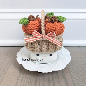 May include: A small crocheted basket filled with orange pumpkins and a red and white gingham bow sits on top of a white ceramic marshmallow with a smiley face and the text "handmadejoy".
