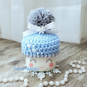 May include: A white ceramic mug with a smiling face, wearing a blue crocheted hat with a gray pom-pom and white snowflakes. The hat has a white ribbon with a snowflake bow. The mug is sitting on a wooden surface with white beads and snowflakes.