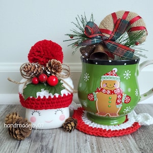 May include: A festive Christmas-themed mug and marshmallow decoration. The green mug features a gingerbread man design and snowflake accents. The marshmallow is adorned with a red pom-pom hat, pinecones, and berries. A red and white crocheted coaster completes the scene.