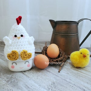 May include: White crocheted chick mug cozy with yellow feet and a red comb. The cozy is sitting on a white mug with a painted face. There are two brown eggs and a yellow chick in a nest behind the mug.
