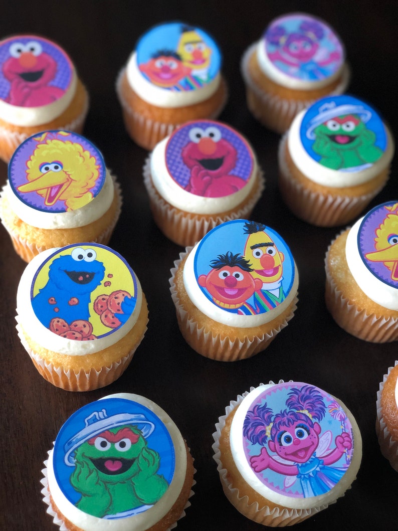 May include: A collection of cupcakes decorated with edible images of Sesame Street characters. The cupcakes are frosted with white frosting and have a variety of colorful images, including Big Bird, Elmo, Cookie Monster, and Abby Cadabby.