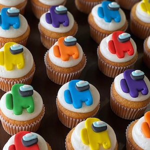 May include: A collection of cupcakes decorated with colorful fondant figures resembling the characters from the popular video game Among Us. The cupcakes are arranged on a brown wooden surface.