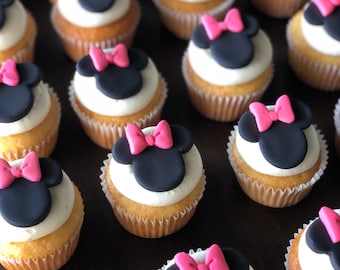 Op de afbeelding: Een close-up van cupcakes versierd met zwart en roze fondant in de vorm van Minnie Mouse's hoofd. De cupcakes zijn in rijen gerangschikt op een donkerbruin oppervlak.
