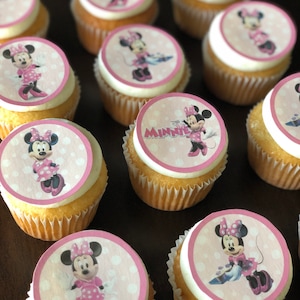 Op de afbeelding: Een close-up van cupcakes versierd met eetbare afbeeldingen met een Minnie Mouse-thema. De cupcakes zijn bedekt met witte frosting en hebben roze en witte cupcake-vormpjes met stippen. De cupcakes zijn gerangschikt op een donkerbruin oppervlak.