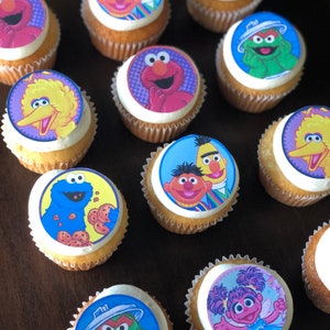 May include: A collection of 11 cupcakes decorated with Sesame Street character images. The cupcakes are frosted with white frosting and have a variety of colorful images of characters like Big Bird, Elmo, Cookie Monster, and Bert and Ernie.