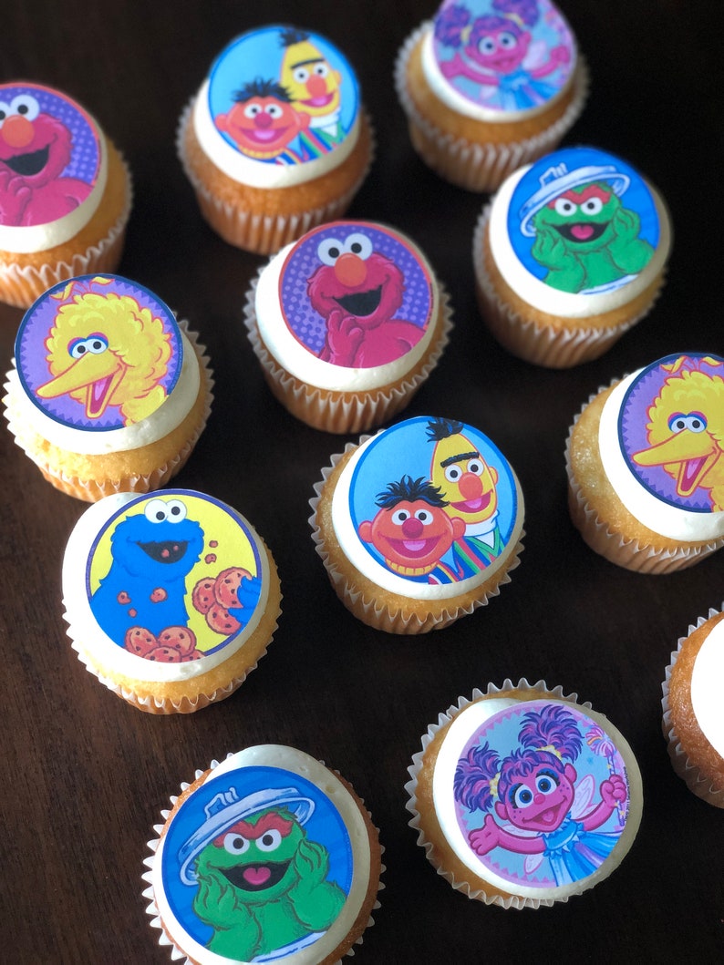 May include: A collection of 11 cupcakes decorated with Sesame Street character images. The cupcakes are frosted with white frosting and have a variety of colorful images of characters like Big Bird, Elmo, Cookie Monster, and Bert and Ernie.