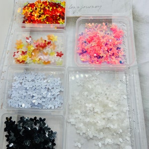 Peut inclure: Six petits contenants en plastique transparent remplis de paillettes en forme de fleurs de différentes couleurs. Les contenants sont disposés en deux rangées de trois. Les couleurs sont le rouge, l'orange, le jaune, le rose, le blanc et le noir.