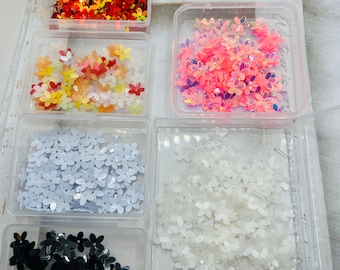 May include: Six small clear plastic containers filled with various colors of flower-shaped sequins. The containers are arranged in two rows of three. The colors are red, orange, yellow, pink, white, and black.