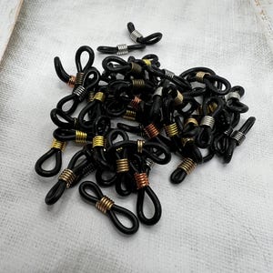 May include: A pile of black eyeglass cord end tips with gold, silver, and copper metal accents. The tips are designed to be attached to eyeglass cords or chains.