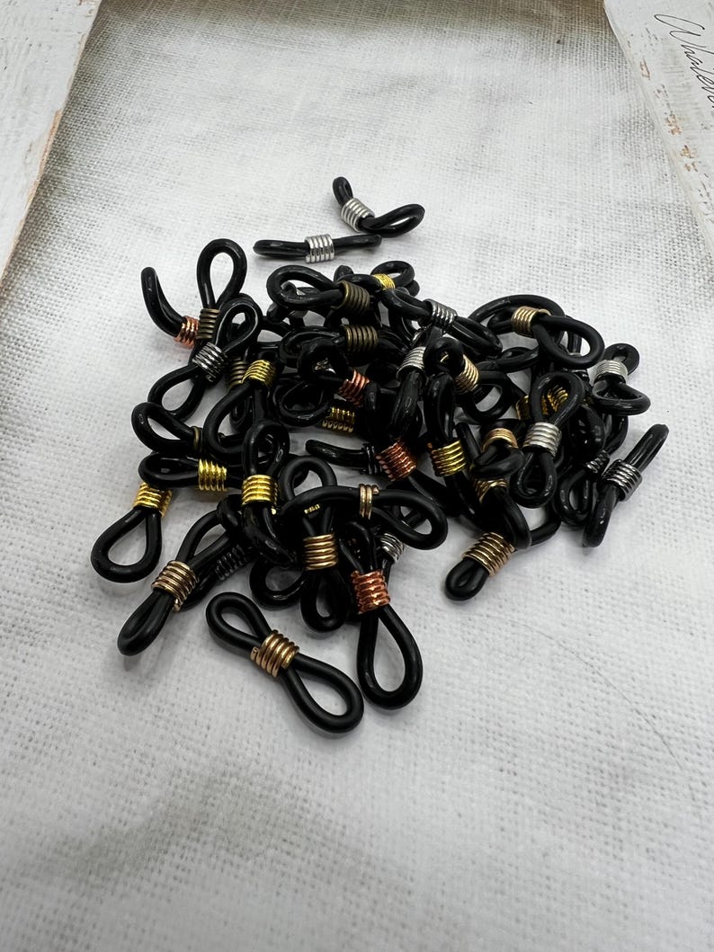 May include: A pile of black eyeglass cord end tips with gold, silver, and copper metal accents. The tips are designed to be attached to eyeglass cords or chains.