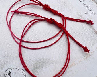 Könnte beinhalten: Zwei rote Armbänder aus Schnur mit Knoten an den Enden. Die Armbänder sind miteinander verbunden und liegen auf einer weißen Oberfläche.