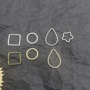 May include: A set of six metal jewellery making blanks in two different finishes. There are two square, two round, and two teardrop shapes. One set is silver and the other is gold.