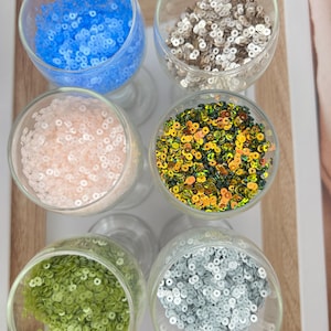 May include: Six clear stemmed glasses filled with colourful sequins. The sequins are in shades of blue, silver, pink, gold, green, and grey. The glasses are arranged on a wooden tray, creating a vibrant display of craft supplies.