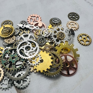 Op de afbeelding: Een verzameling van verschillende metalen tandwielen in verschillende maten en kleuren, waaronder goud, zilver, koper en zwart. De tandwielen zijn willekeurig gerangschikt op een wit oppervlak.
