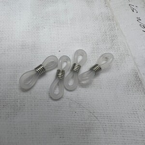 May include: Four clear plastic eyeglass cord holders with silver metal rings. The holders are designed to attach to the ends of eyeglass arms.