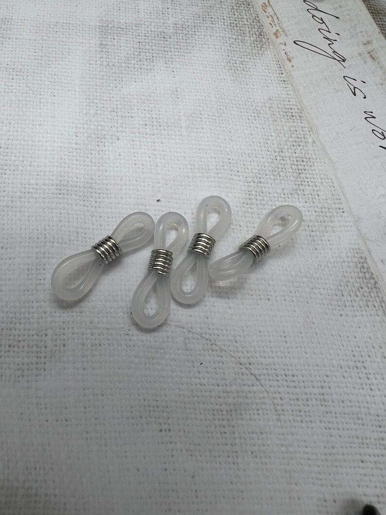 May include: Four clear plastic eyeglass cord holders with silver metal rings. The holders are designed to attach to the ends of eyeglass arms.