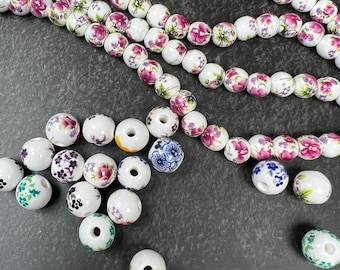 May include: A collection of round white ceramic beads featuring various floral patterns in shades of pink, purple, blue, green, and black. Some beads are strung together on a thin wire, while others are scattered individually, each with a center hole.
