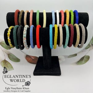 Könnte beinhalten: Eine Präsentation von Perlenarmbändern in verschiedenen Farben, darunter Rot, Grün und Blau, auf einem schwarzen Samtständer. Die Armbänder sind in Reihen angeordnet und zeigen eine Vielzahl von Designs und Farbtönen. Das Bild enthält den Text "Eglantine's World".