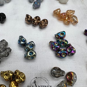 Puede incluir: Surtido de cuentas de calavera de vidrio iridiscente en varios colores, incluyendo transparente, negro, dorado y arcoíris. Las cuentas están dispuestas sobre una superficie blanca con el texto "EGLANTINE'S WORLD" visible en el centro inferior de la imagen.