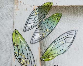 May include: Four green and white cicada wings with gold glitter accents. The wings are arranged in a semi-circle on a white wooden surface. The text "Life is a lot" is visible in the top right corner of the image. The number "#1" is visible in the bottom right corner of the image.