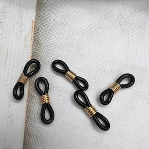 May include: Five black and gold eyeglass cord stoppers. The stoppers are shaped like a figure eight with a gold metal band in the center.