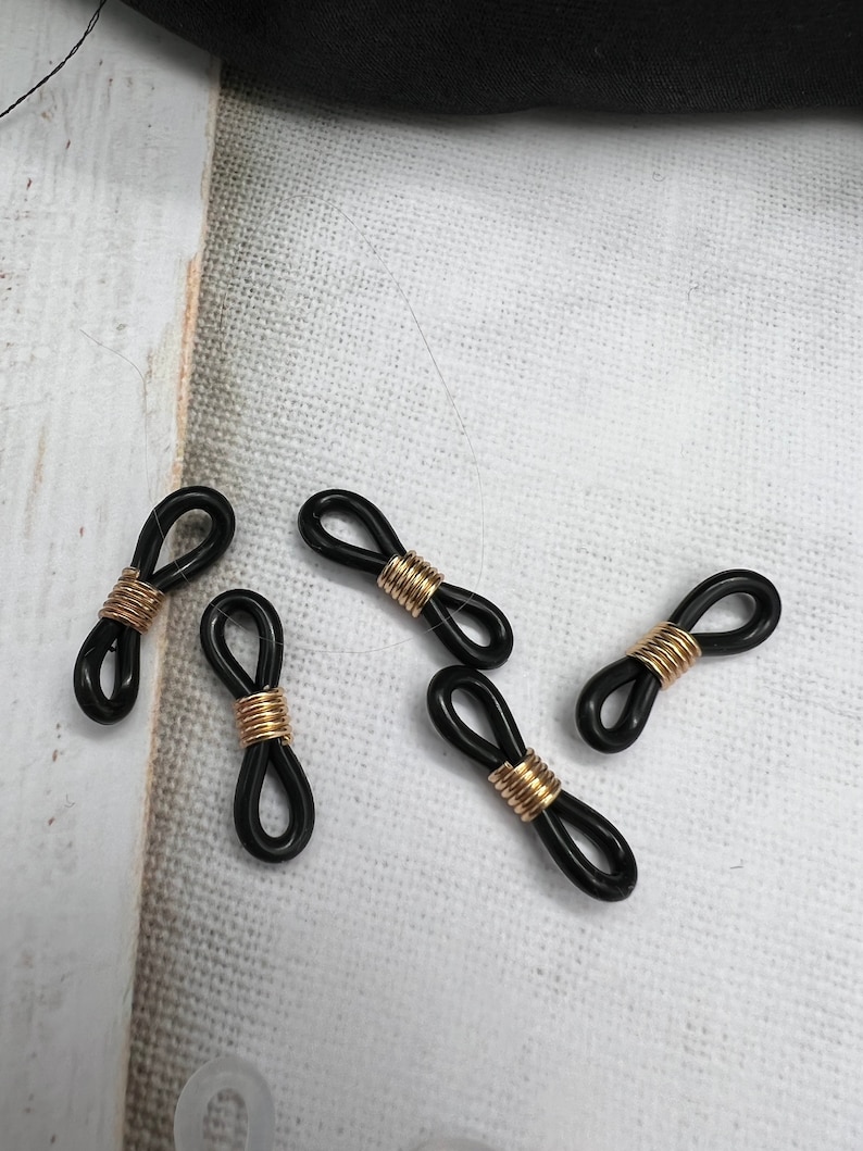 May include: Five black and gold eyeglass cord stoppers. The stoppers are shaped like a figure eight with a gold metal band in the center.