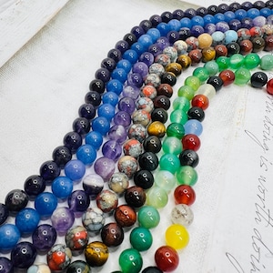 Op de afbeelding: Een verzameling kleurrijke edelsteen kralen in verschillende tinten, waaronder blauw, paars, groen en rood. De ronde kralen zijn geregen, waardoor een regenboogeffect ontstaat. De afbeelding is tegen een witte achtergrond geplaatst.