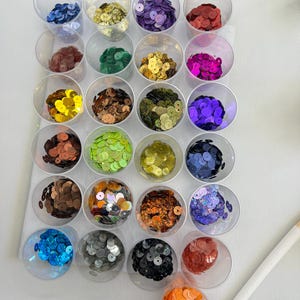 May include: A selection of colourful sequins displayed in small, clear plastic cups. The sequins are in a variety of colours, such as blue, gold, purple, red, brown, green, yellow, and orange. The cups are arranged in a grid pattern on a white surface.