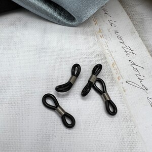 May include: Three black and silver eyeglass holders, also known as eyeglass retainers, are arranged on a white surface. The holders are made of a flexible material and have a spring-loaded design.