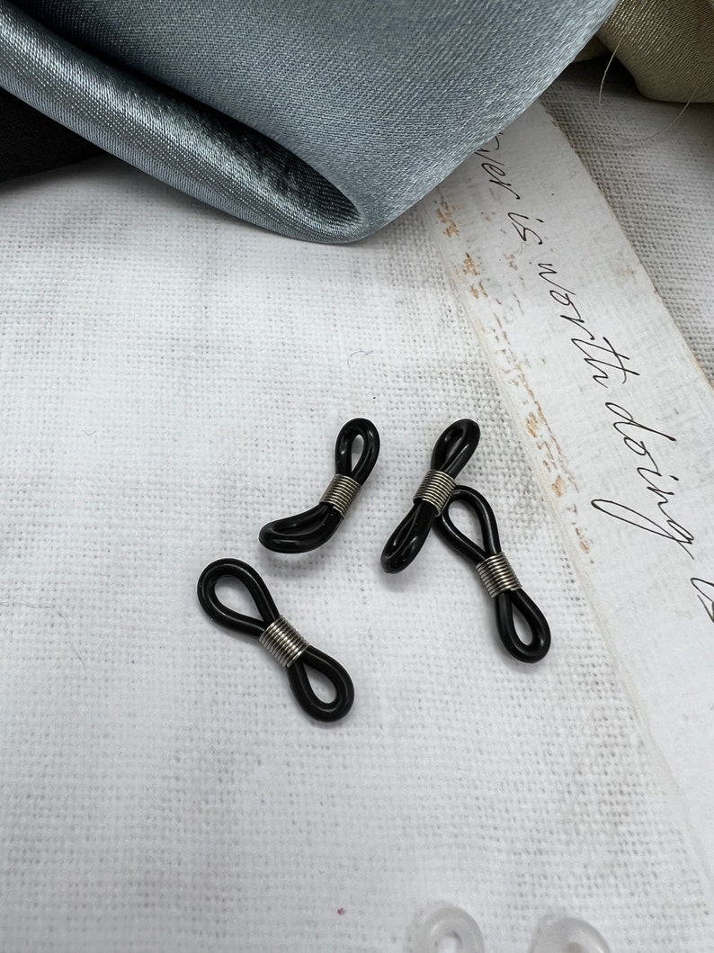May include: Three black and silver eyeglass holders, also known as eyeglass retainers, are arranged on a white surface. The holders are made of a flexible material and have a spring-loaded design.