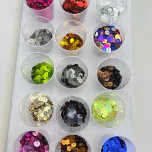 May include: A selection of colourful sequins displayed in clear plastic cups. The sequins are in a variety of shades, such as pink, red, silver, gold, green, black, and blue. The sequins are round and shiny, ideal for crafting and embellishments.