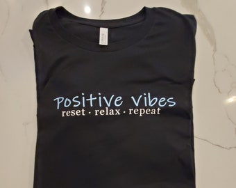 May include: Black t-shirt with the text "Positive vibes reset. relax. repeat" in blue and pink lettering.