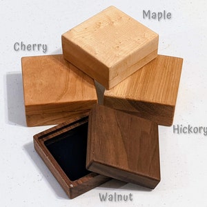May include: Four square wooden boxes labeled Maple, Cherry, Hickory, and Walnut. The boxes are made of different wood tones ranging from light to dark. One dark walnut box is open, revealing a black interior lining, while the others are closed.