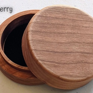 May include: A round wooden box with a natural grain finish and a removable lid. The interior is lined with black fabric. The word Cherry is printed in the upper left corner of the image. The box features a small circular metal magnetic closure.