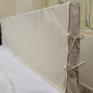 May include: A rectangular headboard cover made of off-white fabric. The side panel is a contrasting beige color and features two sets of fabric ties that are knotted to secure the cover in place along the side edge of the headboard structure.