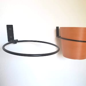 May include: Two black metal plant hangers mounted on a white wall. One hanger has a terracotta pot with a plant inside. The other hanger has a ring for a pot.