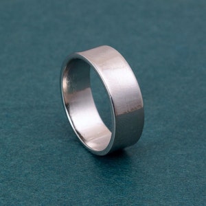 May include: A silver-colored ring with a flat, wide band. The ring has a brushed metal texture and a simple, modern design. The ring is shown on a teal background. Jewelry.
