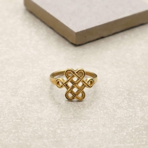 Puede incluir: Un anillo dorado con un intrincado diseño de nudo. El anillo presenta un patrón central tejido con elementos en forma de corazón y acentos circulares. El anillo se muestra sobre una superficie gris claro.