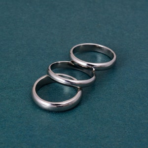 May include: Three silver rings are arranged in a curved line against a teal background. The rings are simple bands with a polished finish, reflecting light. The rings are likely for a wedding or engagement.